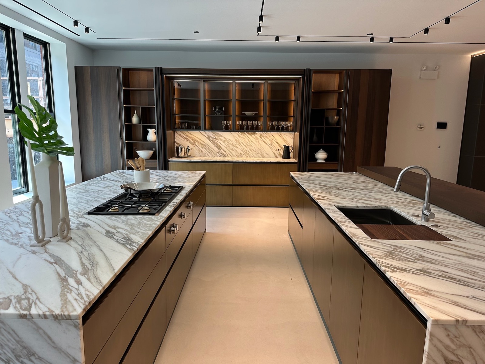 Two working kitchen islands in marble and bronze before a lit glass cabinet wall
