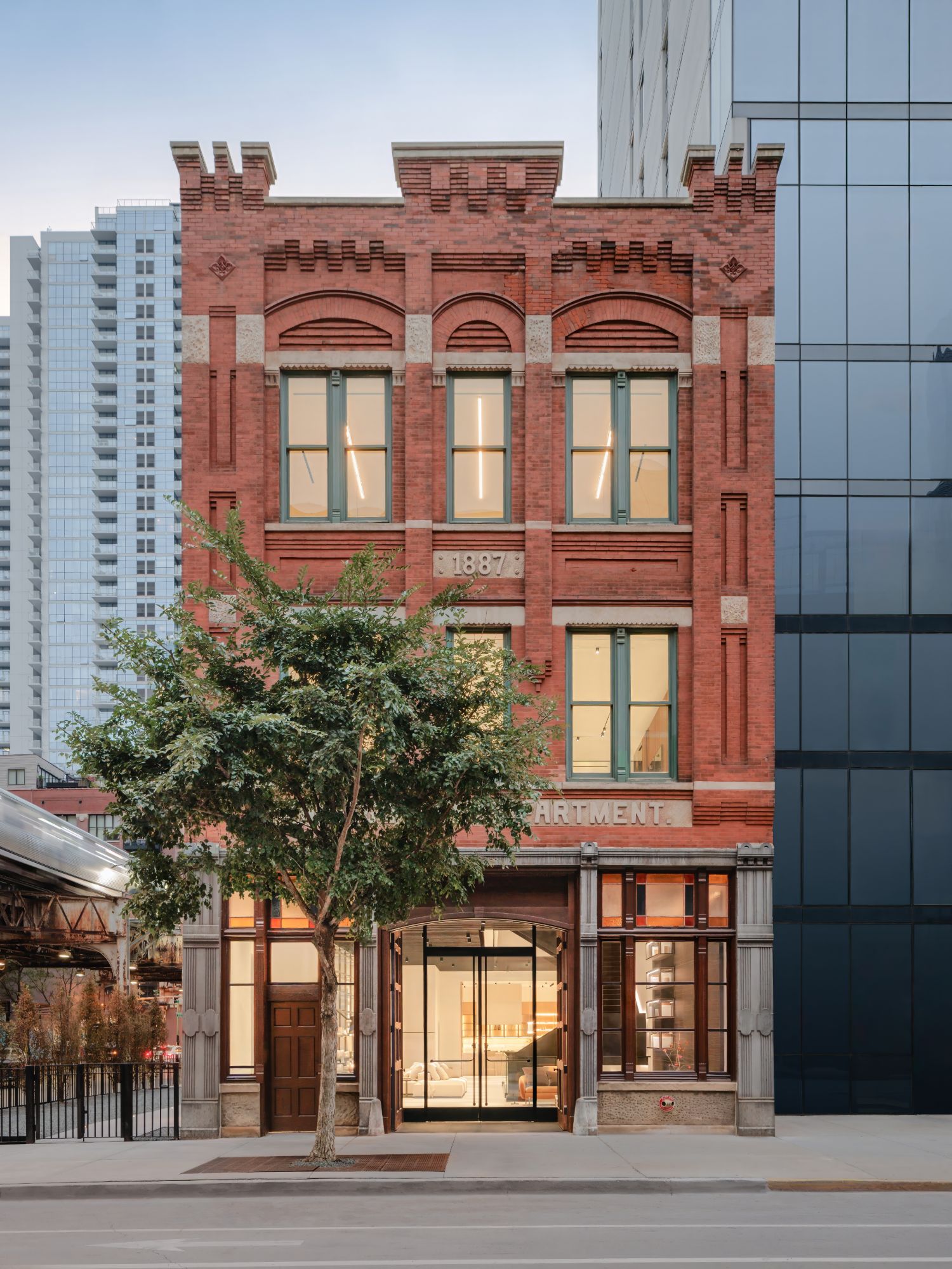 The restored 1887 firehouse at dusk, seen whole from its crenellated roofline to the sidewalk, between Chicago towers